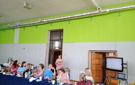 Sala gimnastyczna, w tle telewizor z prezentacją o Erasmus oraz nauczyciele siedzący przy stole