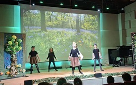 zdjęcie przedstawia scenę w sali koncertowej. Na pierwszym planie, od strony widowni, widać głowy siedzących dzieci oglądających występ. Na scenie stoi grupa dzieci przebranych w proste stroje &mdash; czarne bluzki i brązowe sp&oacute;dniczki lub spodnie. Niekt&oacute;re maj