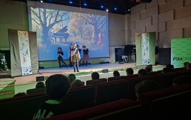 drugie zdjęcie przedstawia scenę w sali koncertowej. Na pierwszym planie, od strony widowni, widać głowy siedzących dzieci oglądających występ. Na scenie stoi grupa dzieci przebranych w proste stroje &mdash; czarne bluzki i brązowe sp&oacute;dniczki lub spodnie. Niekt