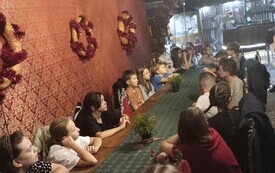 Na zdjęciu dzieci siedzą przy stole i czekają na wskaz&oacute;wki od pani prowadzącej warsztaty