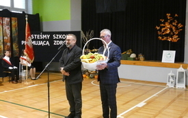 Naczelnik oświaty oraz zastępca prezydenta miasta stoją na środku sali, jeden z nich trzyma kosz z owocami