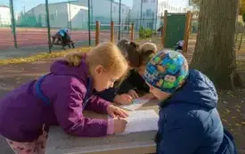 Zdjęcie 3 przedstawi troje dzieci. Dziewczynki w kolorowych kurtkach , uśmiechnięte. Chłopiec pochylony nad oparciem ławki. Na ławce leżą kart pracy, kt&oacute;re dzieci wypełniają, realizując projekty matematyczny.
