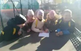 Uczniowie w kolorowych czapkach i kurtkach pochylają się nad kartami pracy z zadaniami matematycznymi. Wsp&oacute;łpracują i są zainteresowani. Zdjęcie zrobiono na placu zabaw.