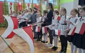 Uczniowie ustawieniu w dwurzędzie, śpiewają pieśń i machają flagami