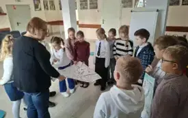 Bibliotekarka ogląda i omawia razem z dzićmi, kt&oacute;re stoją w kole, ich wykonane plakaty dotyczące odzyskania Niepodległości. Dzieci są skupoone na swoich pracach, a Bibliotekarka razem z dziewczynką trzyma w dłoniach duży karton papieru na kt&oacute;rym są symbol