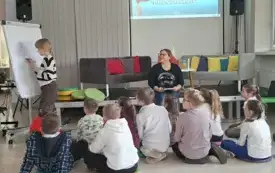 Zdjęcie przedstawiają grupę dzieci siedzących na podłodze i słuchających siedządzej na wprost nich Pani bibliotekarki, kt&oacute;ra trzyma w rękach książkę. Pani prowadzi zajęcia dotyczące ochodzonego już święta Narodowego Dnia Niepodległości. Na ścianie wisi pr