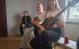Troje uczni&oacute;w siedzi na ławce, jedna uczennica trzyma w rękach gitarę