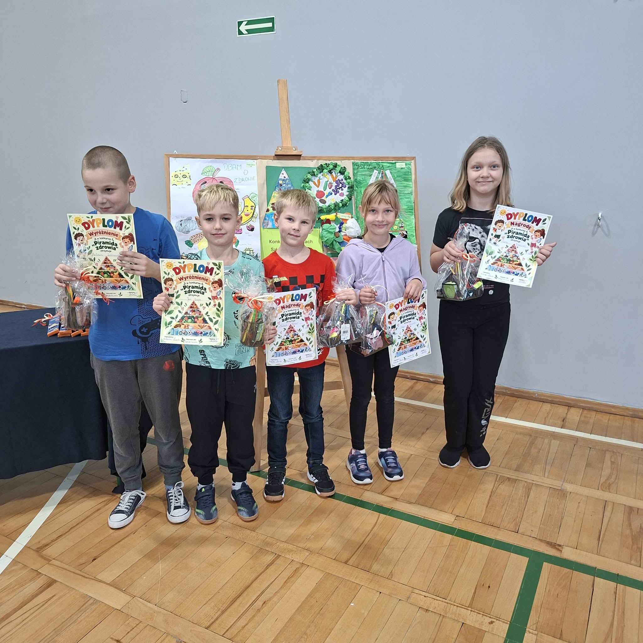 Zdjęcie przedstawia uśmiechnięte dzieci z klas młodszych, stojące przed tablicą korkową, na której wyeksponowano konkursowe prace plastyczne. Dzieci trzymają w rękach nagrody i dyplomy.