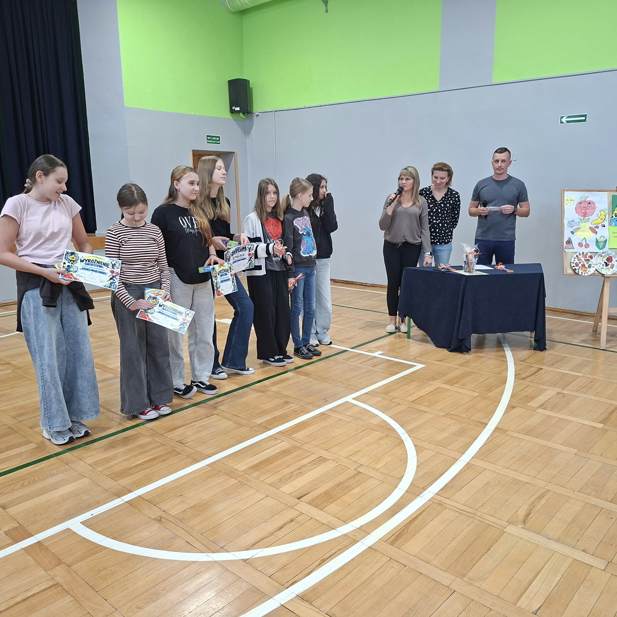 Zdjęcie przedstawia uczniów klas starszych, stojących w szeregu na sali gimnastycznej i trzymających otrzymane nagrody. Po prawej stronie, przy granatowym stoliku nakrytym granatową tkaniną, stoją organizatorzy konkursu – dwie nauczycielki i nauczyciel