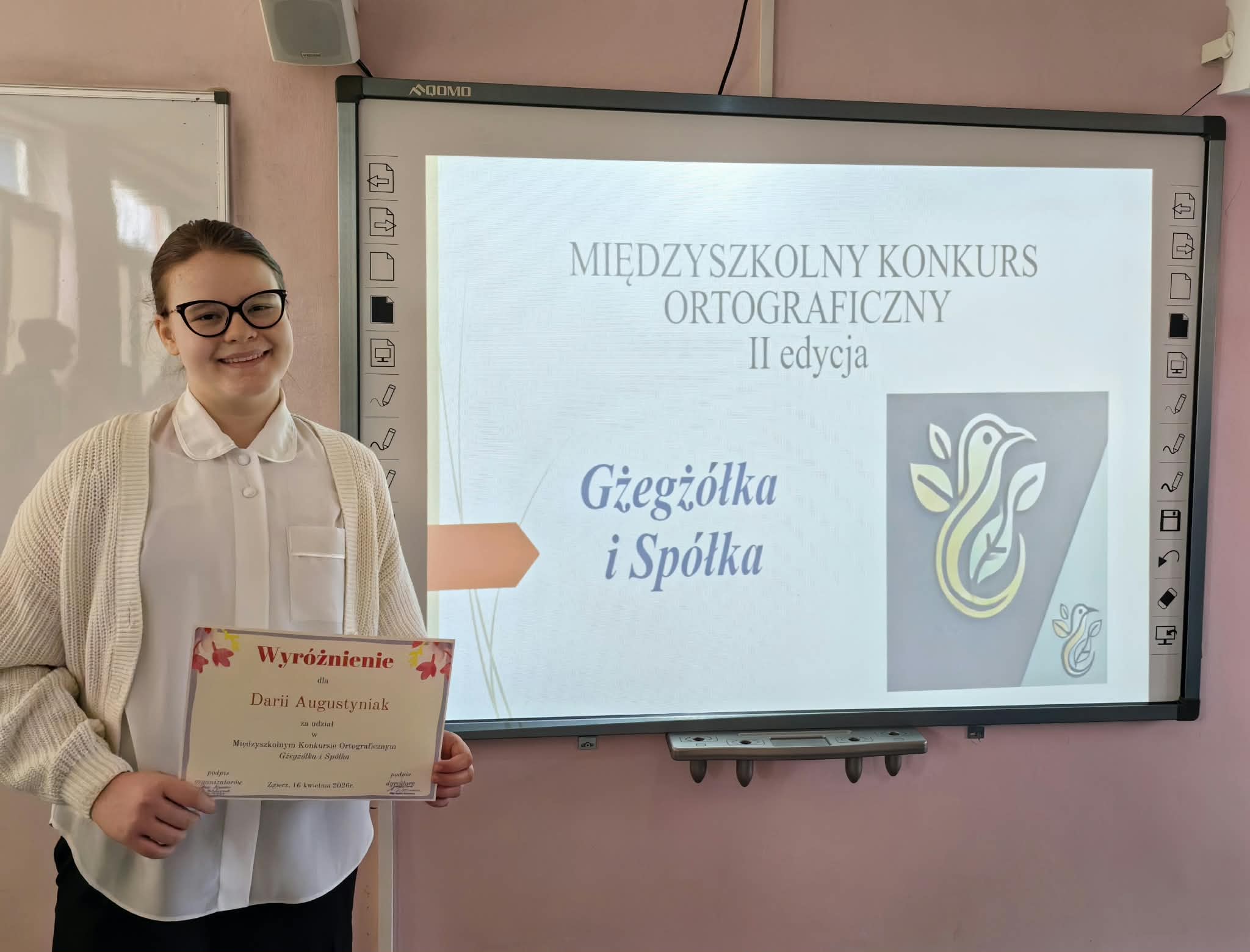 Uczennica pozuje z&nbsp;dyplomem, stoi przy tablicy multimedialnej