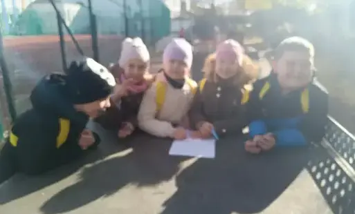 Uczniowie w kolorowych czapkach i kurtkach pochylają się nad kartami pracy z zadaniami matematycznymi. Współpracują i są zainteresowani. Zdjęcie zrobiono na placu zabaw.
