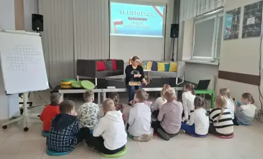 Zdjęcie przedstawiają grupę dzieci siedzących na podłodze i słuchających siedządzej na wprost nich Pani bibliotekarki, która trzyma w rękach książkę. Pani prowadzi zajęcia dotyczące ochodzonego już święta Narodowego Dnia Niepodległości. Na ścianie wisi pr