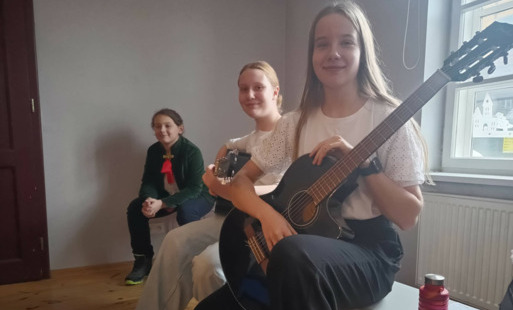 Troje uczniów siedzi na ławce, jedna uczennica trzyma w rękach gitarę
