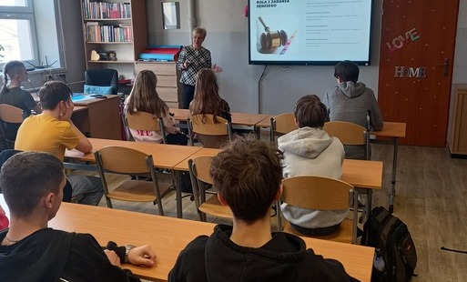 Uczniowie siedzą w ławkach i słuchają wypowiedzi kobiety