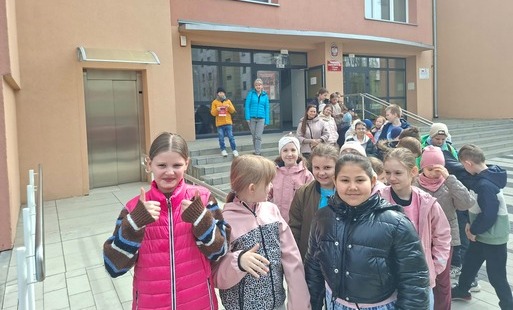 widać grupę dzieci stojących przed budynkiem szkoły muzycznej. Budynek ma jasnobeżowe ściany i duże przeszklone drzwi wejściowe, do których prowadzą schody z metalową poręczą. Nad wejściem znajduje się tabliczka z godłem Polski oraz napisem wskazującym na