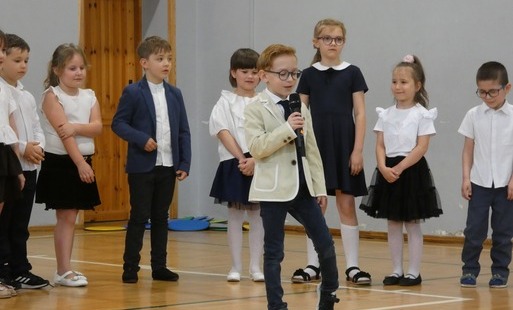jedno dziecko stoi z przodu sceny i przemawia przez mikrofon, pozostałe z tyłu